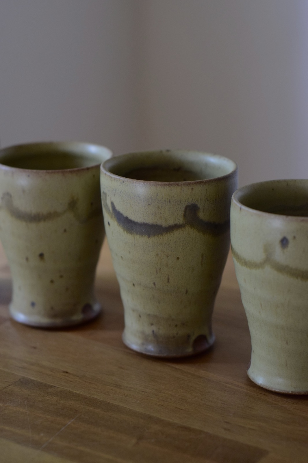 Three stoneware tumblers with an olive ash glaze on a wooden table